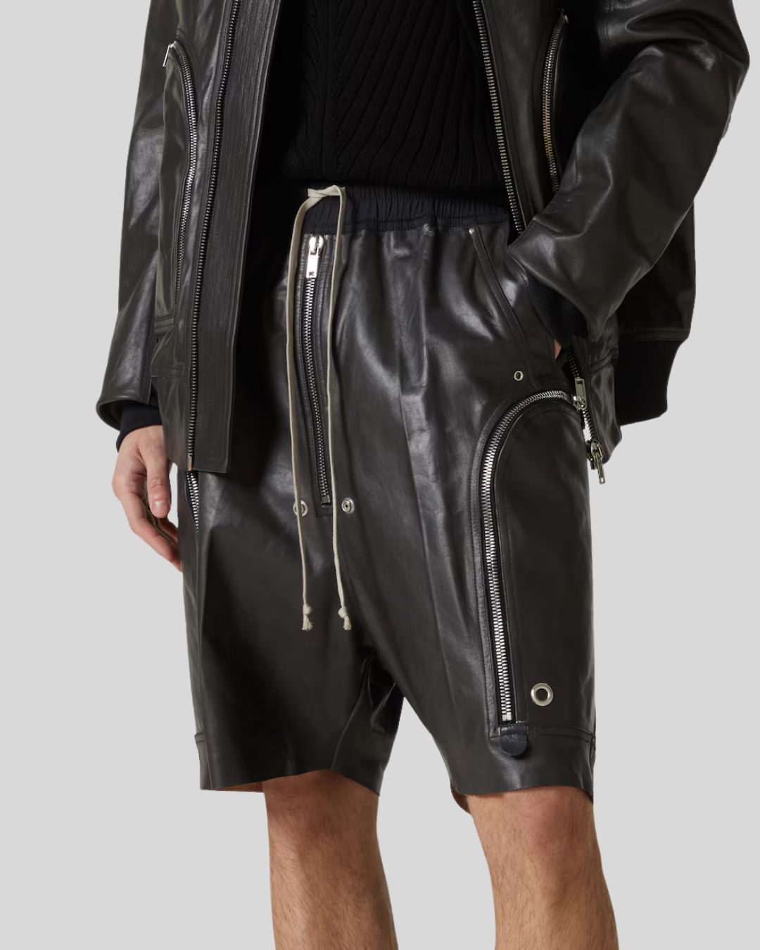 Person wearing black leather shorts with zippers and a black jacket on a light gray background