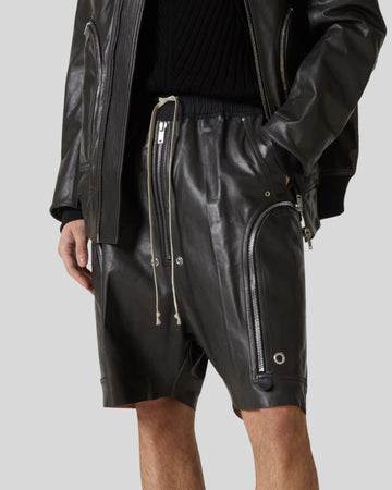Person wearing black leather shorts with zippers and a black jacket on a light gray background